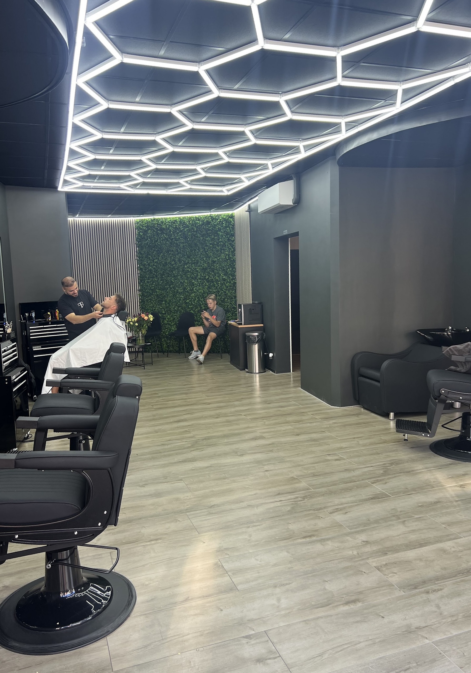 Binnen bij Niko’s Barbershop: zwarte barbierstoelen onder het zeshoekige lichtplafond, achterin wordt een klant geknipt